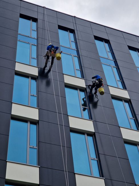 Two professional window cleaners wearing safety harnesses and helmets are suspended on ropes outside a modern high-rise building with blue-tinted glass windows and dark gray panels. They are using squ