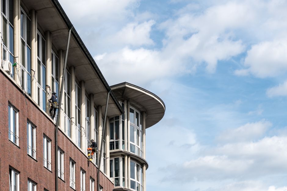 Two professional window cleaners wearing safety harnesses and helmets are suspended on ropes outside a modern high-rise building with blue-tinted glass windows and dark gray panels. They are using squ