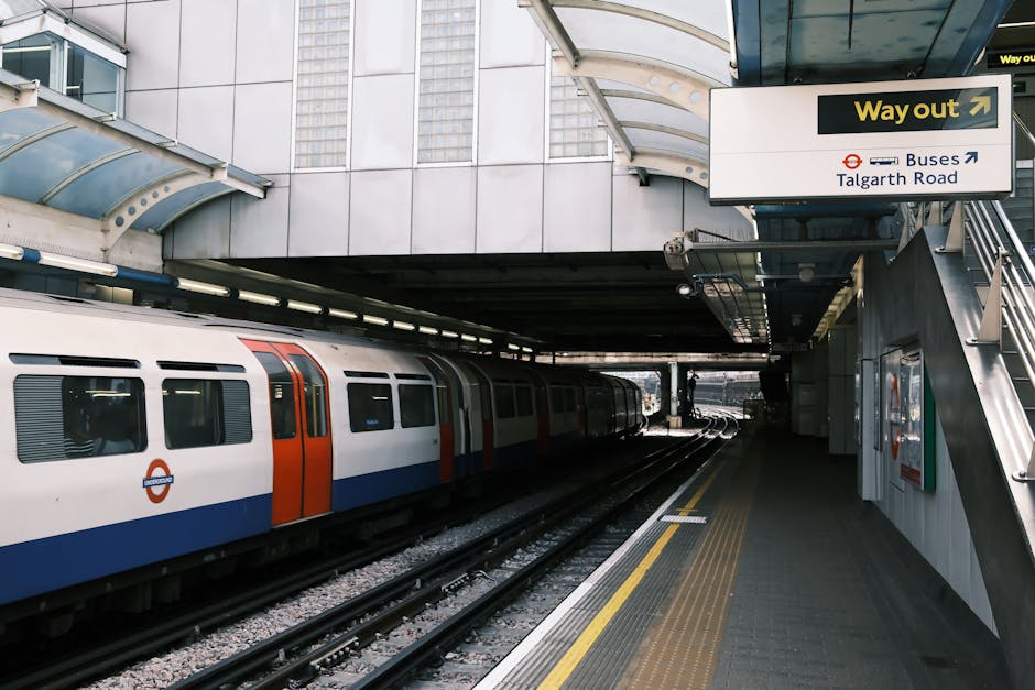 The image depicts a modern train station platform with a sleek, stainless steel train featuring red and blue livery parked along the tracks. The platform surface is made of concrete tiles with tactile
