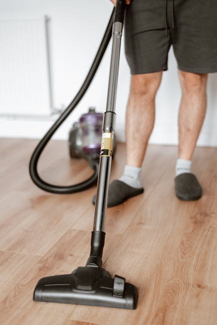 A person wearing grey shorts, grey slippers, and white socks is using a handheld vacuum cleaner to clean a wooden floor in a residential setting. The vacuum has a black hose and a silver and black bod